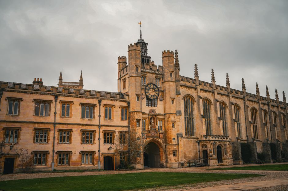 A historic beige stone building with Gothic architectural features, including tall arched windows, decorative battlements, and a central tower with intricate spires, situated behind a well-maintained