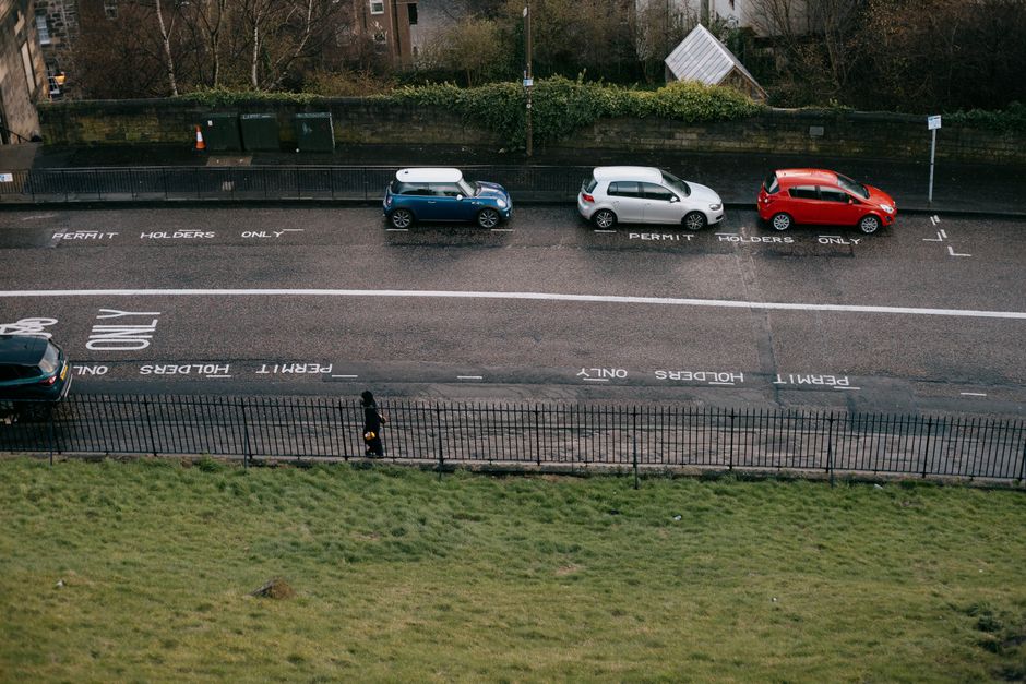 An aerial view of a street showing three parked cars – a blue compact vehicle with a white cargo box on its roof, a white hatchback, and a red hatchback – aligned along a designated permit holder'